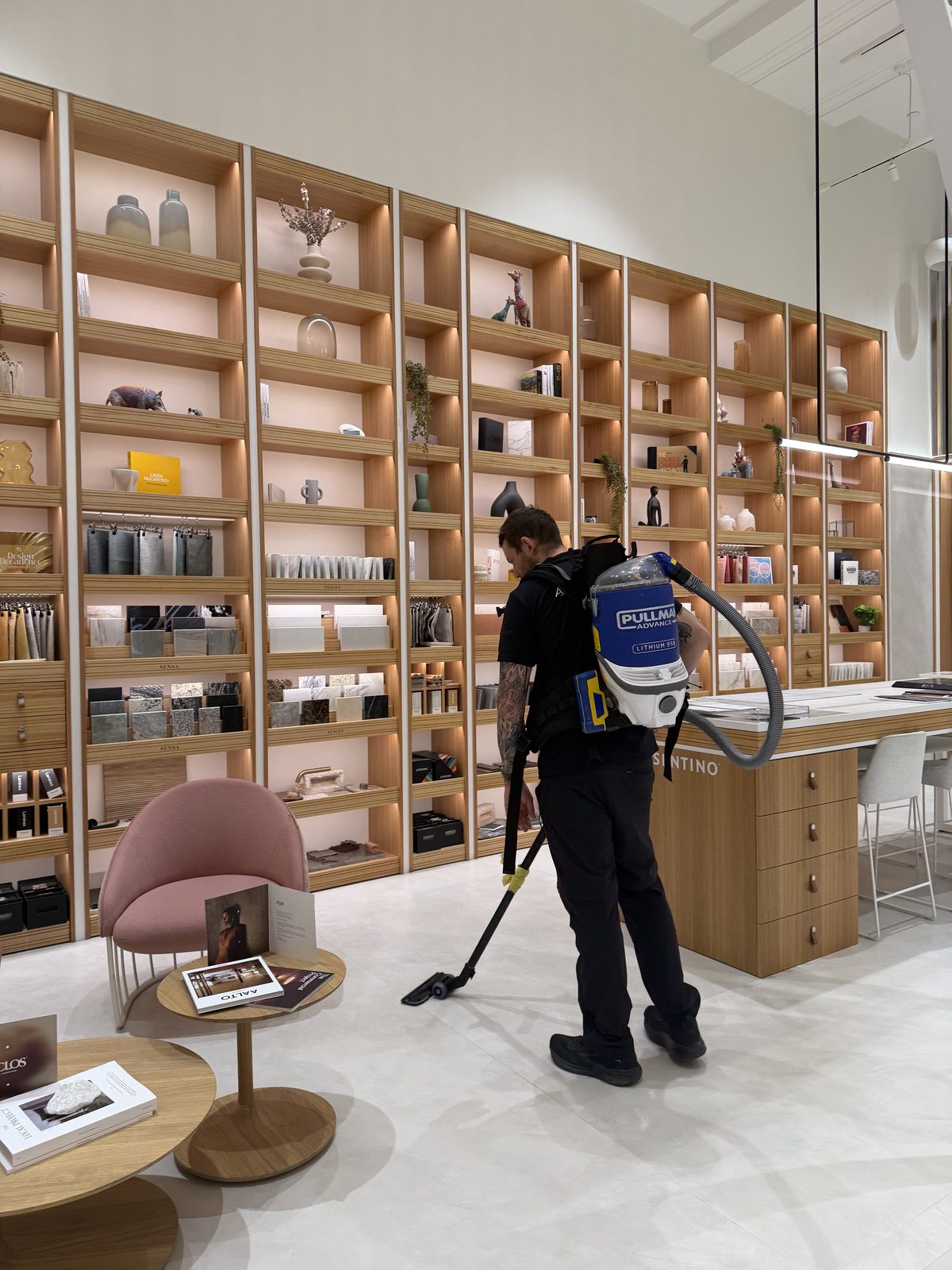 Cleaner wearing a backpack vacuum working along a full-height illuminated display shelving wall of samples and objects in the showroom