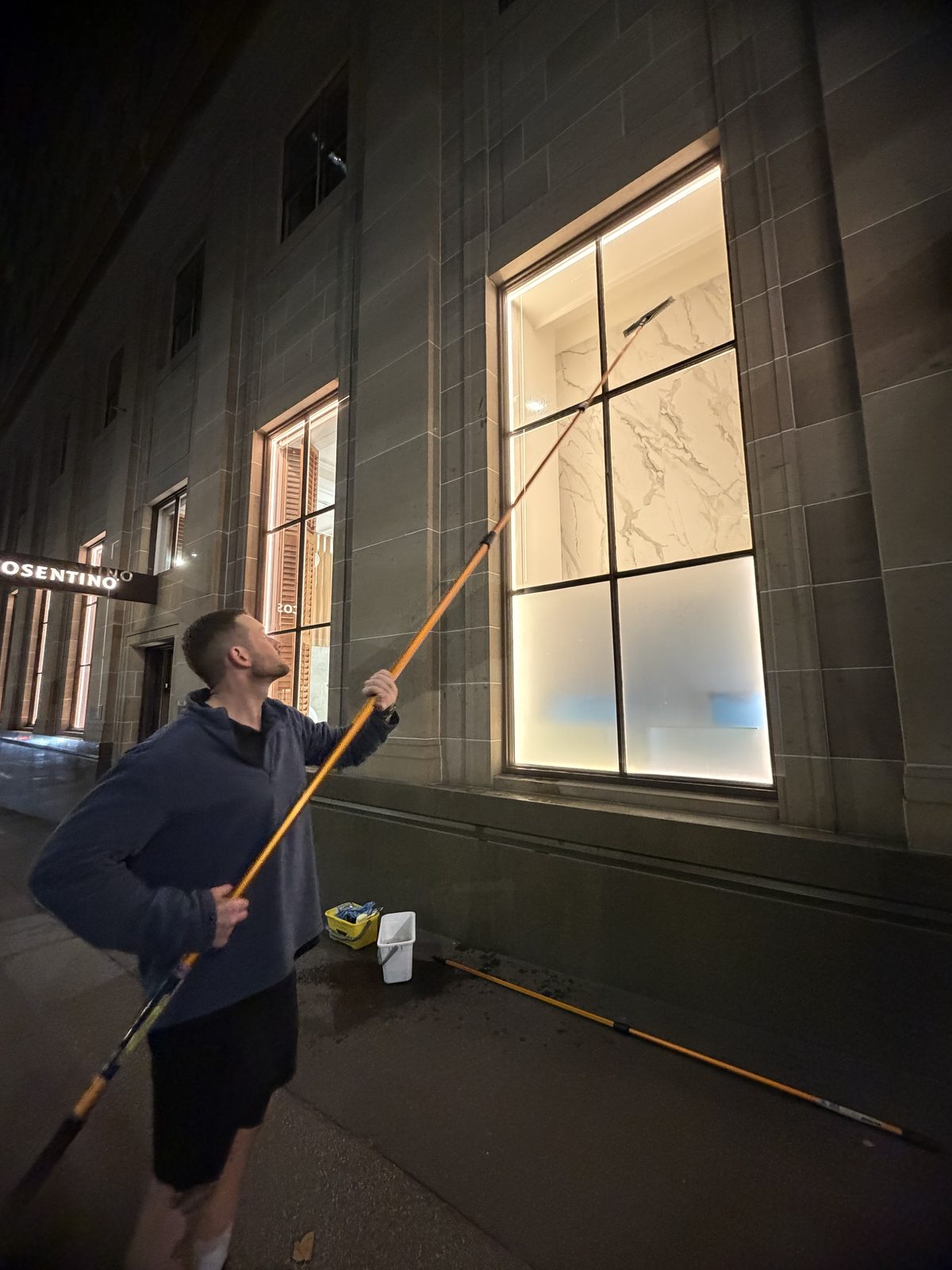 Cleaner washing an exterior showroom window at night with an extension pole, the lit marble-lined interior visible through the glass