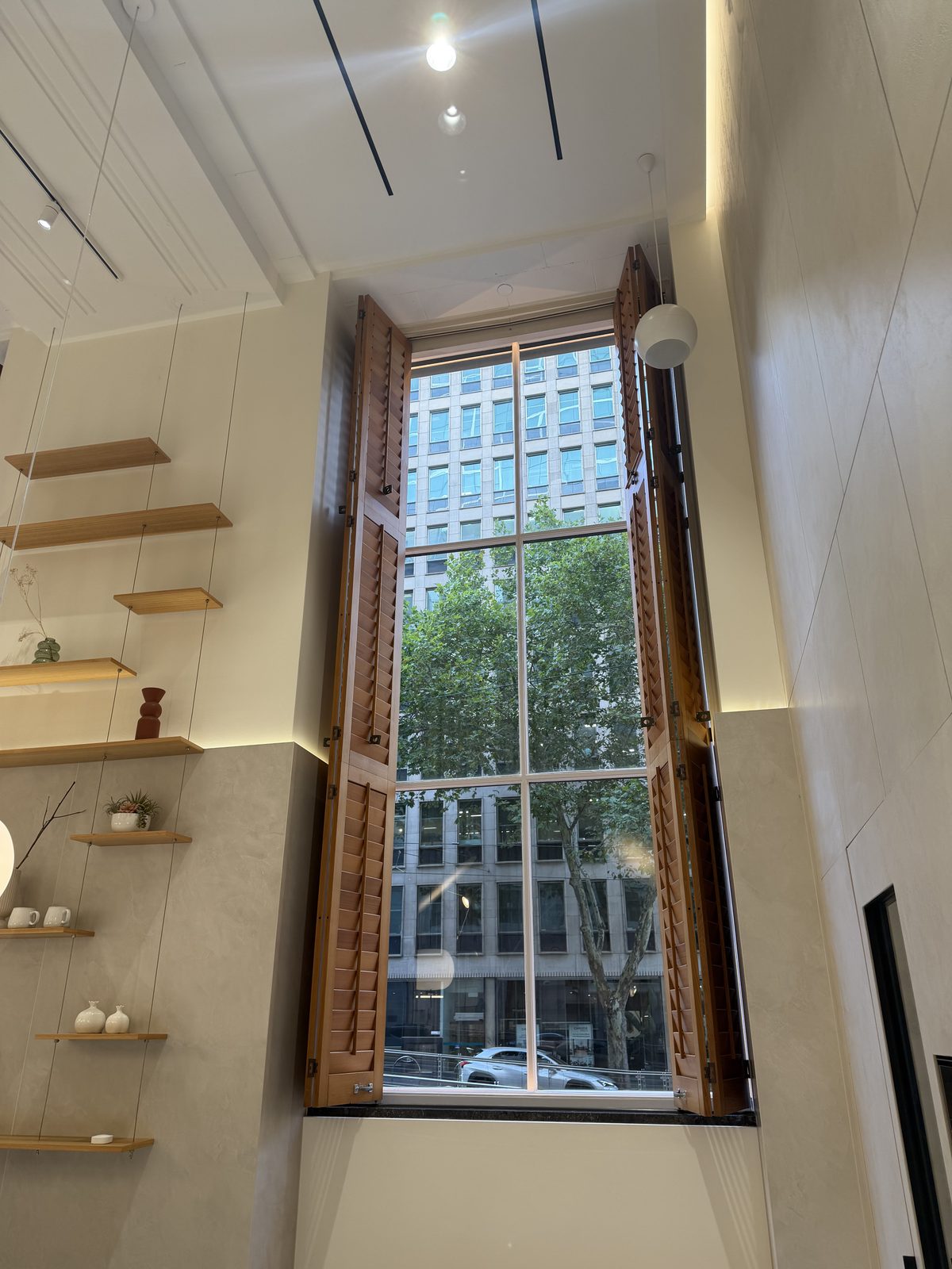 Floor-to-ceiling multi-pane window with open timber shutters, the glass completely clear with street trees and buildings visible outside