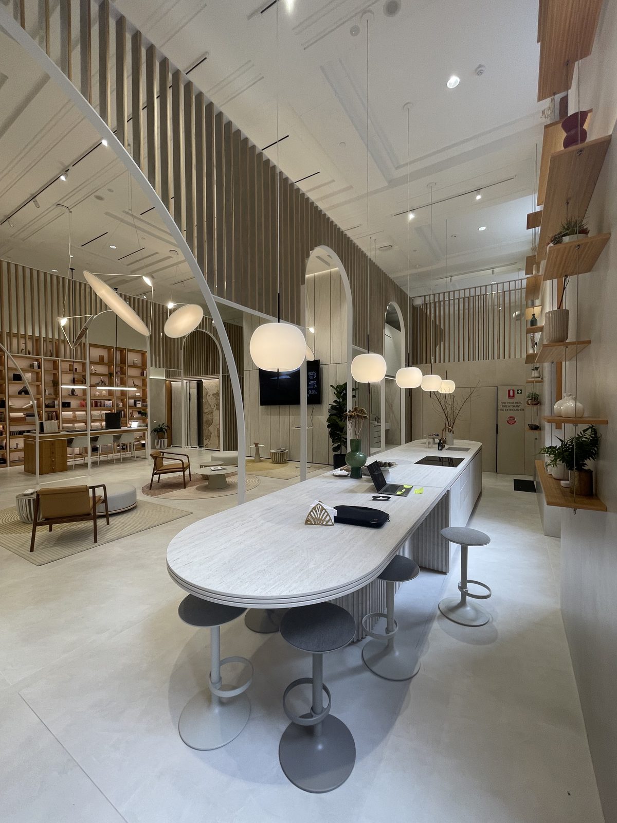 Cosentino showroom interior with a long travertine island bench and stools under pendant lights, timber-batten walls and display shelving beyond, floors and surfaces spotless