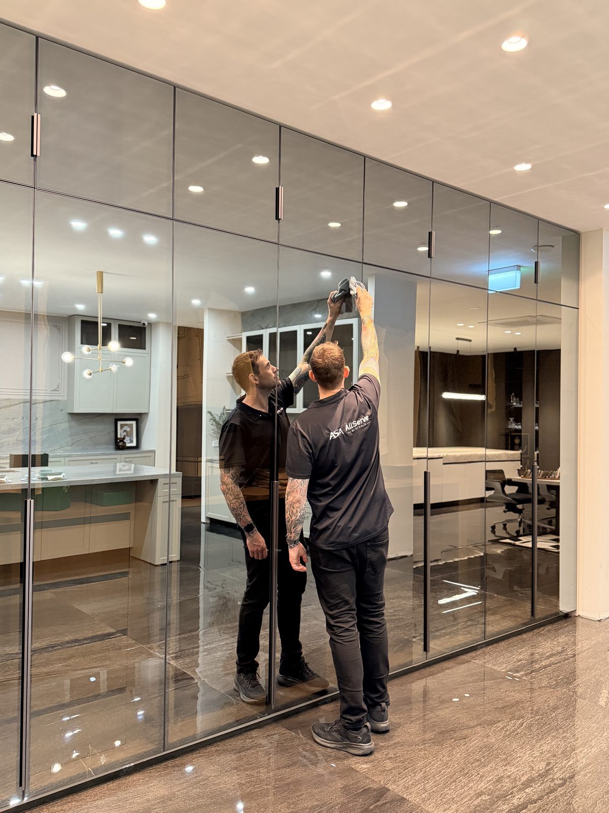 Uniformed cleaner wiping a wall of floor-to-ceiling mirrored glass cabinet doors in the showroom, with the display kitchen reflected in the glass