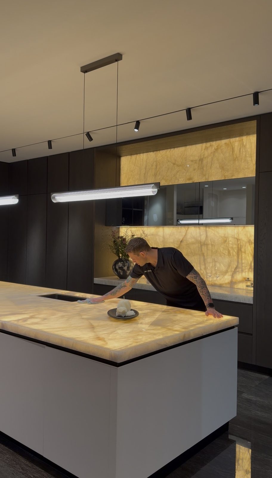 Cleaner wiping down a backlit onyx island benchtop in a dark-toned display kitchen at the Cremorne showroom