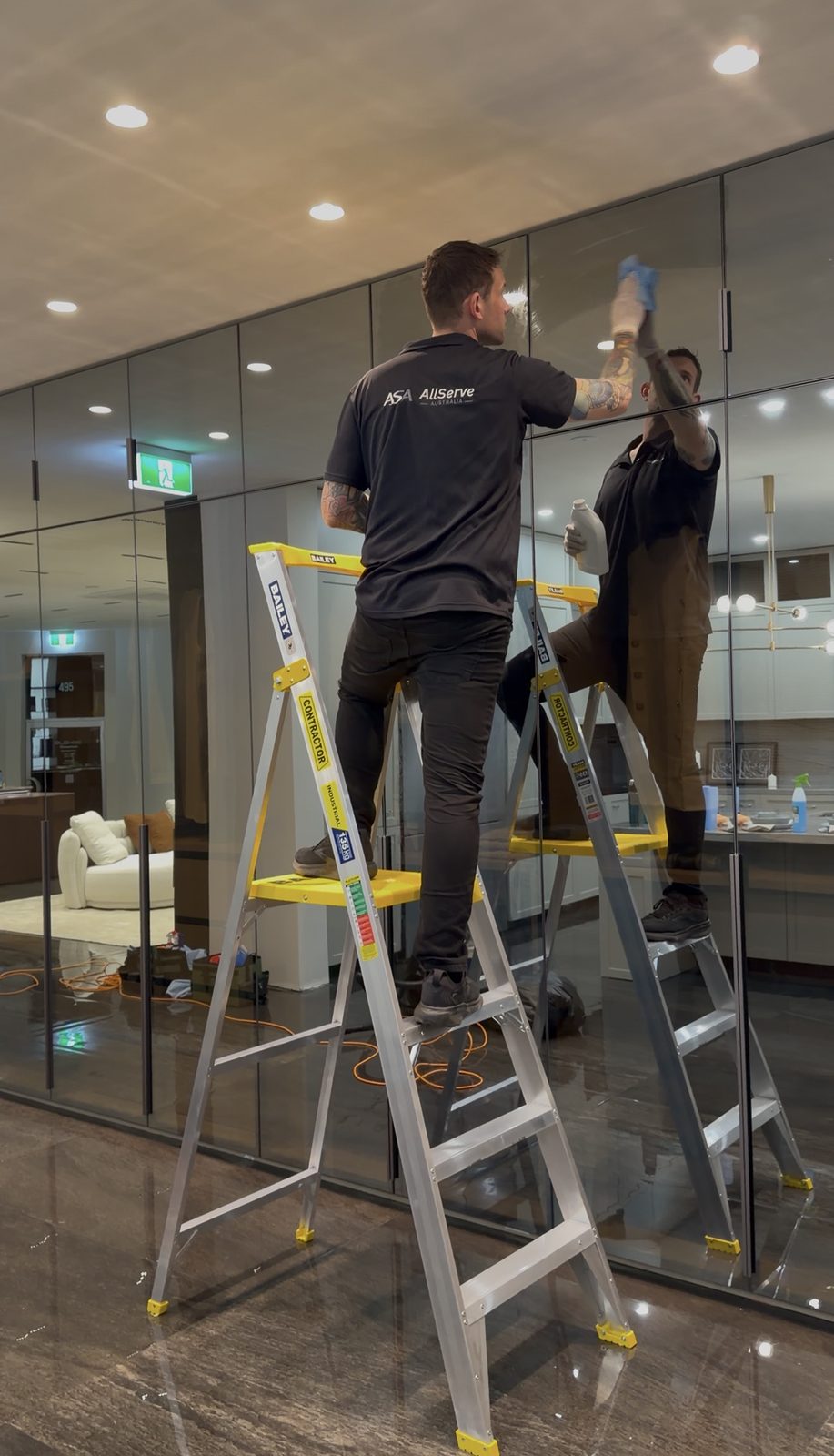 Two cleaners on step ladders cleaning the top edge of a full-height glass cabinet wall in the showroom