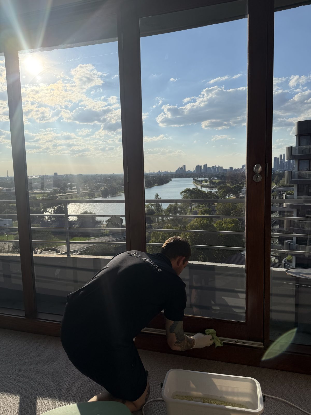 Cleaning technician wiping down the glass and lower frame of a floor-to-ceiling window door in a St Kilda apartment, with a lake and city skyline visible outside.