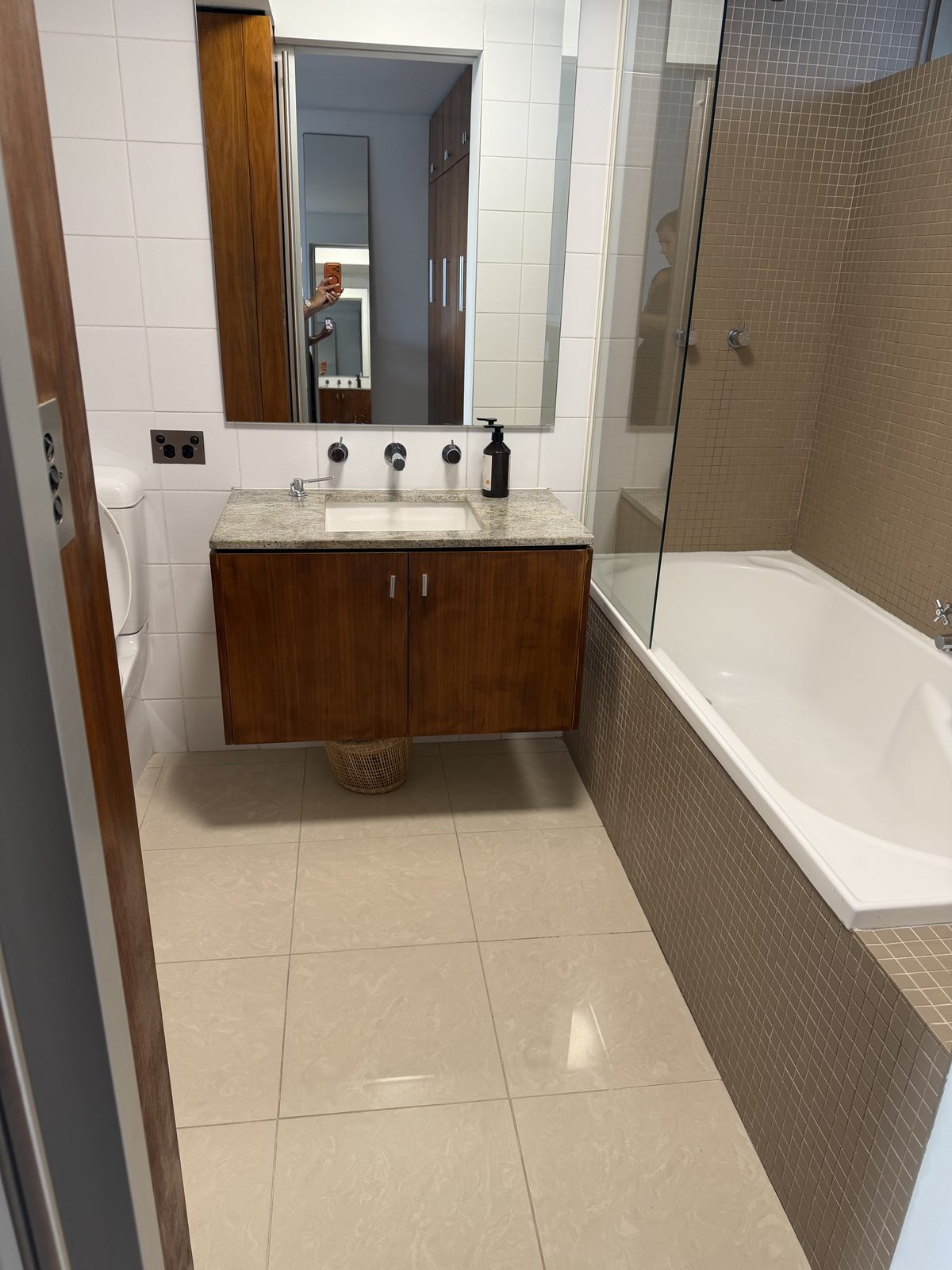 Cleaned bathroom in a St Kilda apartment after an end of lease clean, showing a timber vanity with stone top, mirror, white bathtub with glass screen and polished floor tiles.