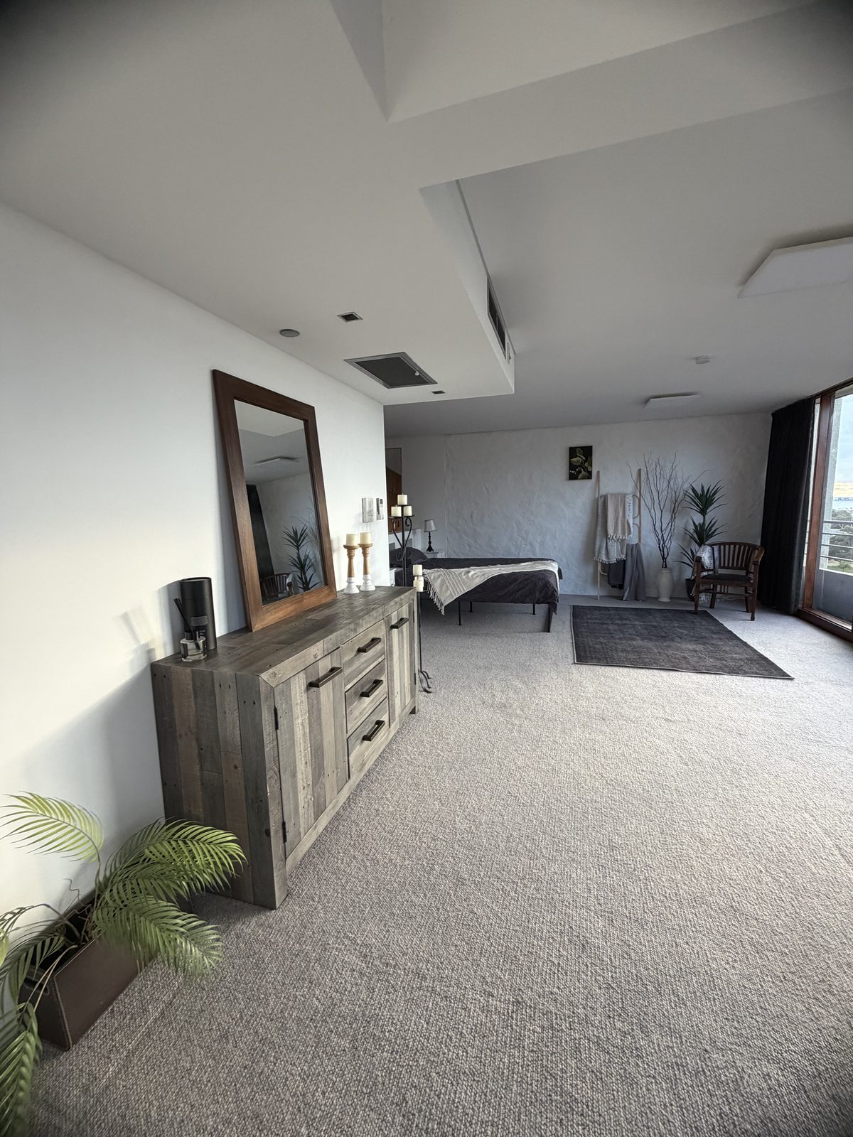 Large carpeted bedroom area of a St Kilda apartment after an end of lease clean, with a mirror and timber dresser, potted plants and a made bed in the background.